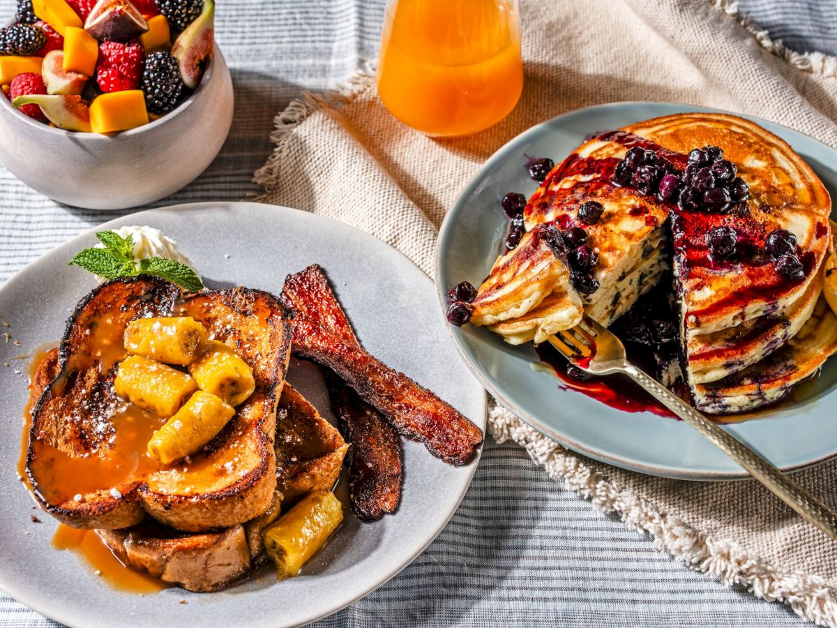 The image shows a breakfast spread with French toast and bananas foster, pancakes with blueberries, a bowl of fresh fruit, and a glass of orange juice.