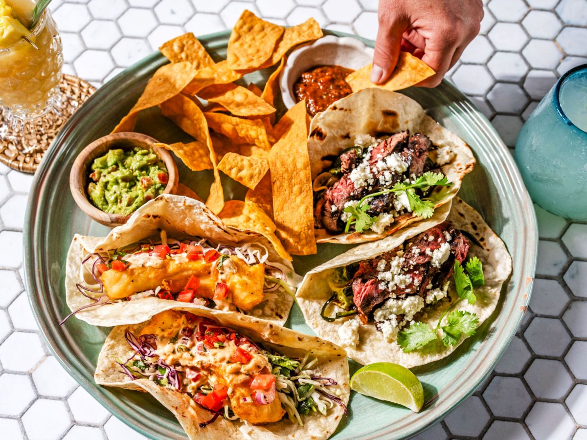 A plate featuring three tacos with various fillings, chips, guacamole, salsa, and beverages like a margarita and a pineapple cocktail are shown.