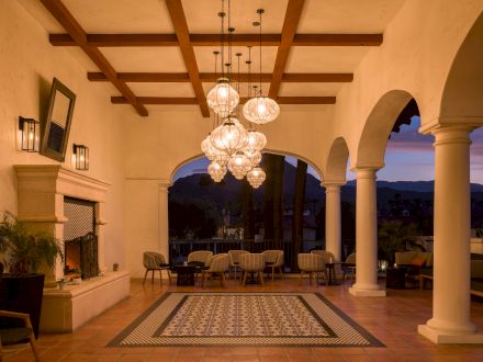 An elegant outdoor patio with chandeliers, wooden beams, arched openings, seating areas, and a fireplace. The dusk sky is visible in the background.