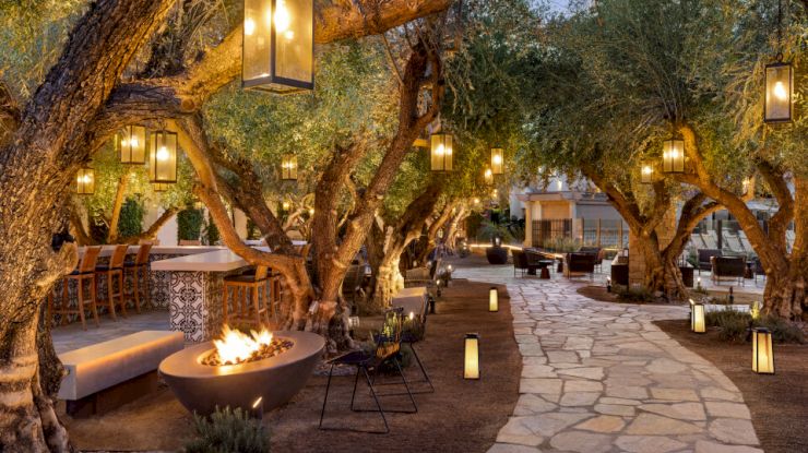 An outdoor dining area with a stone path, hanging lanterns, trees, bar seating, and a fire pit, creating a cozy atmosphere.