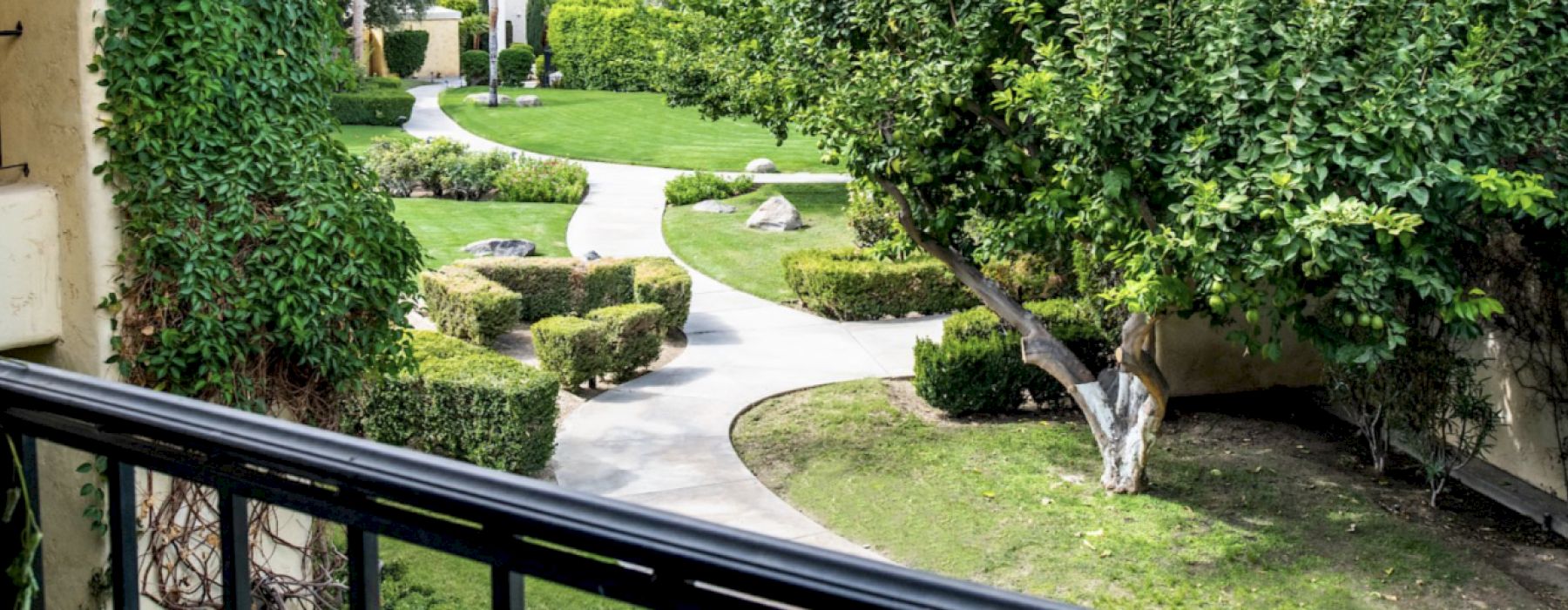 A beautifully landscaped courtyard with a winding pathway, lush greenery, trimmed hedges, and trees, viewed from a balcony with a railing.