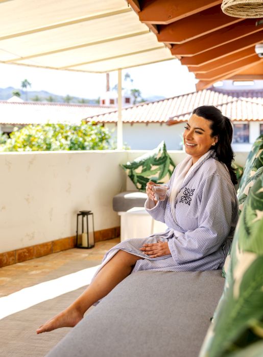 A person is sitting on a patio, wearing a robe and holding a drink, with plants and mountains visible in the background.