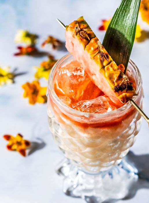A tropical cocktail garnished with a pineapple slice and leaf, served in an ornate glass with scattered flowers around it.
