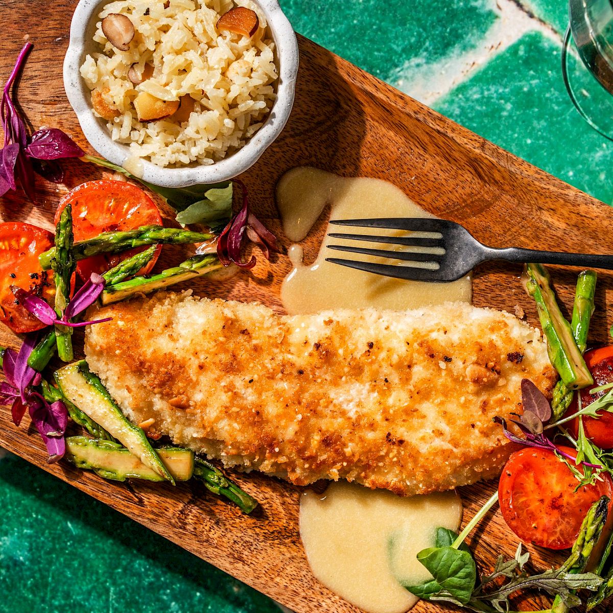 A wooden platter with a breaded fish fillet, assorted vegetables, a small bowl of rice, and a glass of white wine on a green tiled surface.
