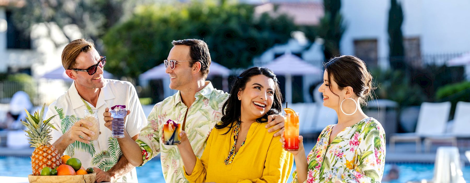 Four people are enjoying drinks by the poolside bar, surrounded by bottles and fresh fruit, with a scenic background of trees and mountains.