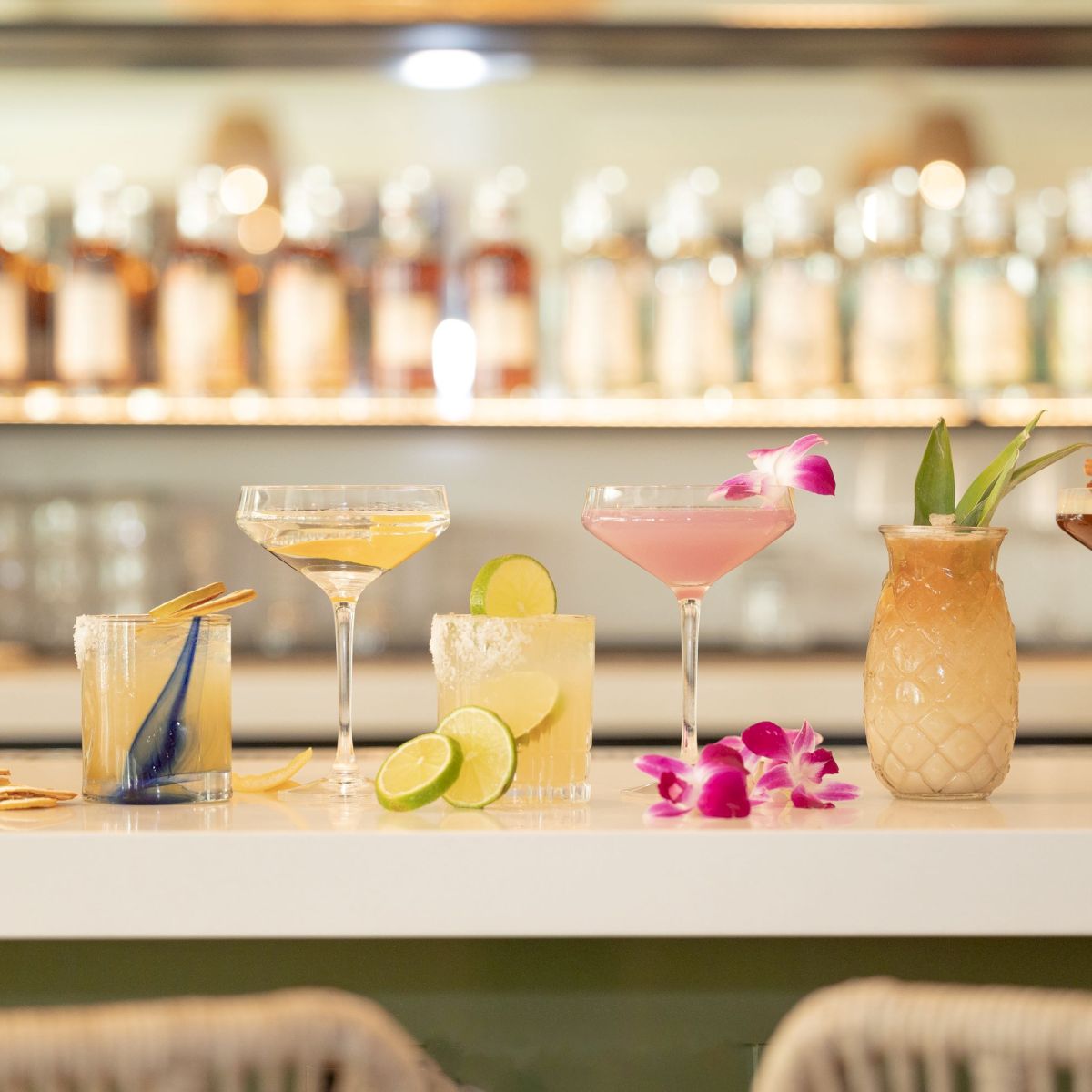 An assortment of cocktails on a bar counter, with garnishes like lime slices, flowers, and a bottle in the background.