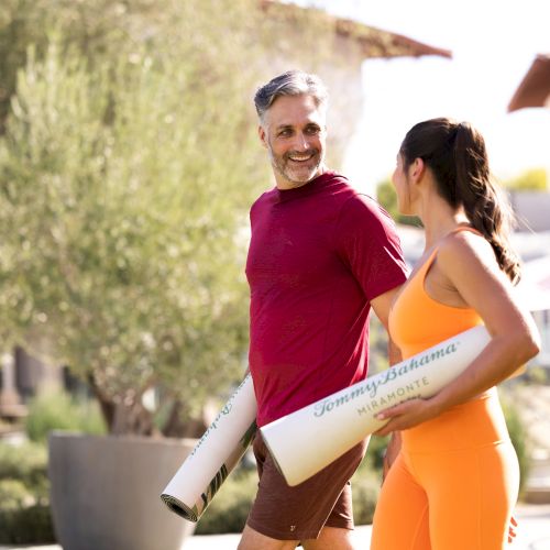 A man in a red shirt and a woman in an orange outfit are walking outside, each carrying a Tommy Bahama Wellness mat.
