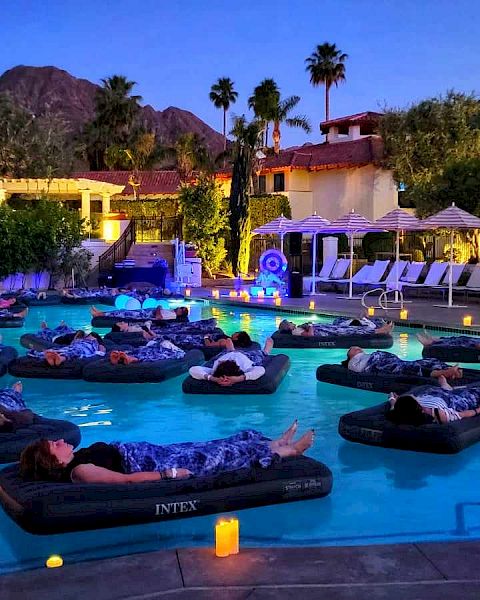 People are relaxing on inflatable loungers in a dimly lit outdoor pool area surrounded by mountains and resort amenities, with candles everywhere.