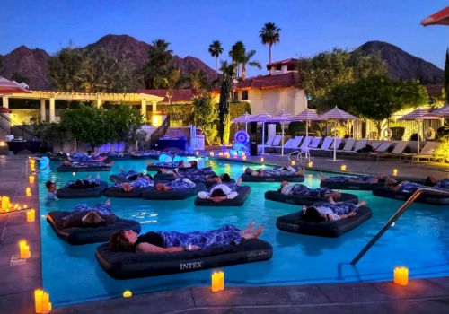People are relaxing on inflatable loungers in a dimly lit outdoor pool area surrounded by mountains and resort amenities, with candles everywhere.