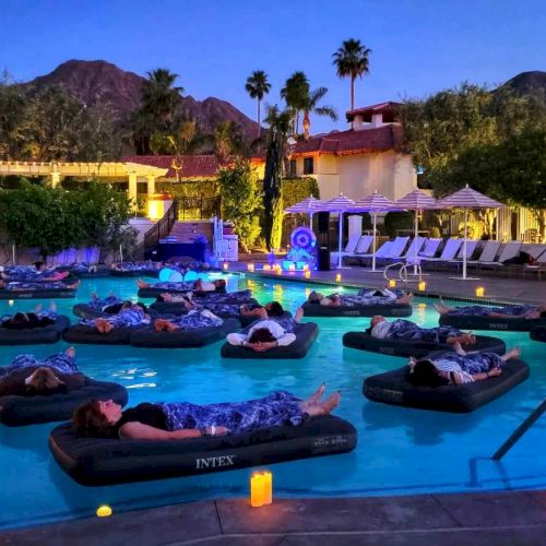 People are relaxing on inflatable loungers in a dimly lit outdoor pool area surrounded by mountains and resort amenities, with candles everywhere.