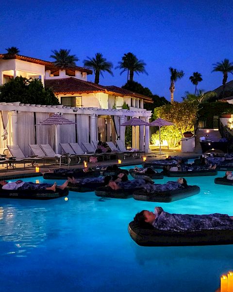 Pool at a resort at night with people floating on inflatable beds, surrounded by lit candles, lounge chairs, and trees in the background.