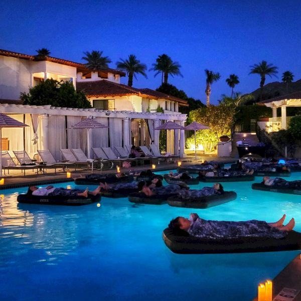 Pool at a resort at night with people floating on inflatable beds, surrounded by lit candles, lounge chairs, and trees in the background.