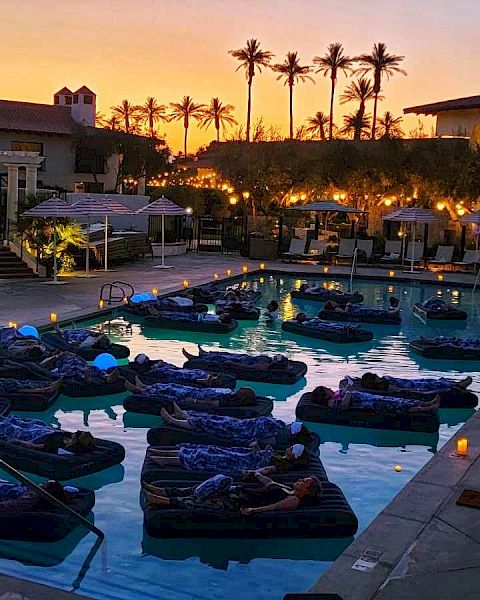 People relax on floating beds in a pool at sunset in a resort, surrounded by loungers, umbrellas, and dim lighting, creating a tranquil ambiance.