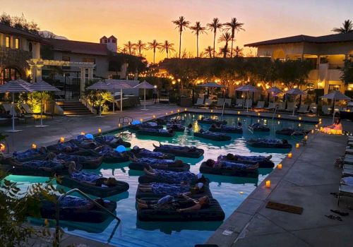 People relax on floating beds in a pool at sunset in a resort, surrounded by loungers, umbrellas, and dim lighting, creating a tranquil ambiance.