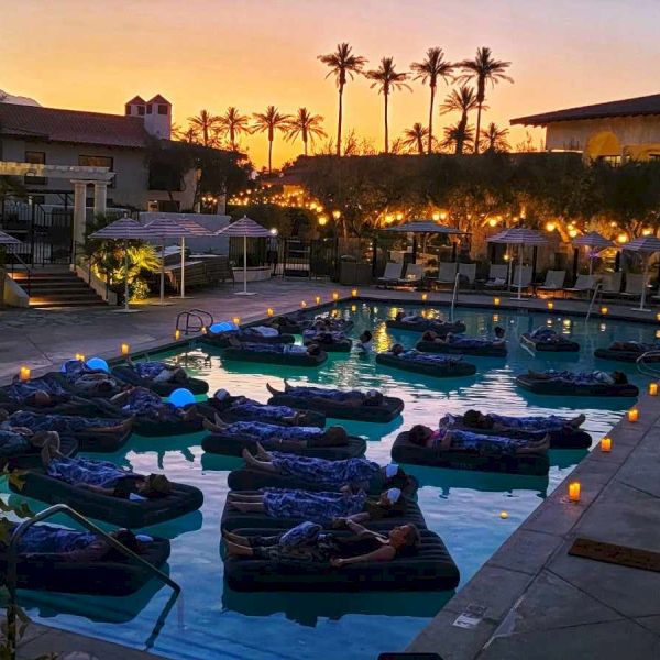 People relax on floating beds in a pool at sunset in a resort, surrounded by loungers, umbrellas, and dim lighting, creating a tranquil ambiance.