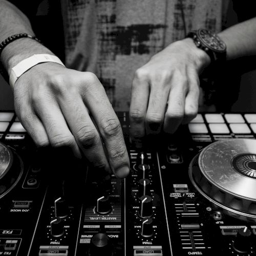 A close-up of a DJ's hands manipulating controls on a DJ mixer, with turntables and various knobs visible in a black and white setting.