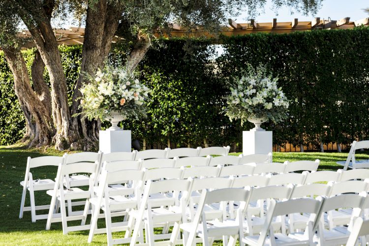 The image shows an outdoor wedding setup with rows of white chairs facing a decorated altar area with two large floral arrangements and trees in the background.