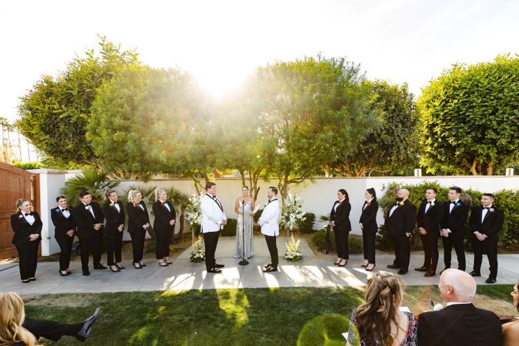 This image shows an outdoor wedding ceremony with a wedding party dressed in black and white attire, standing on either side of the couple.