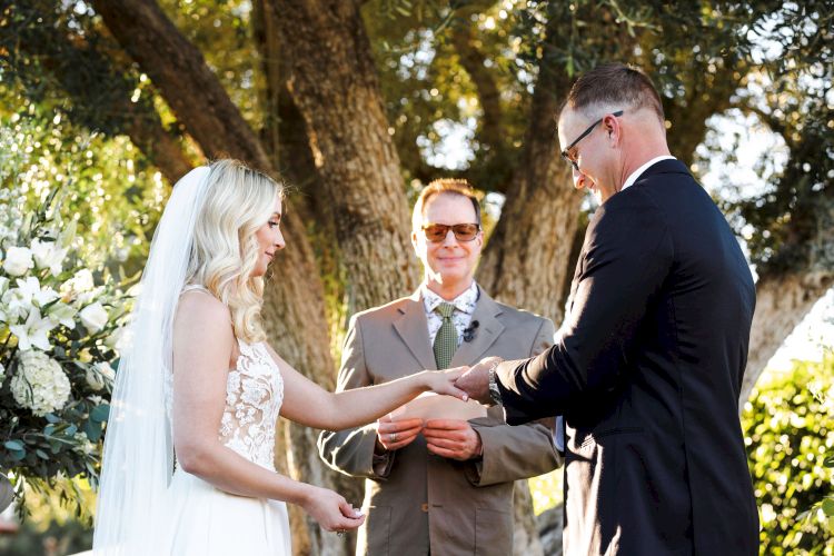 A bride and groom exchange rings during an outdoor wedding ceremony, with an officiant standing behind them.