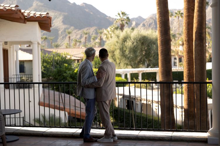 Two individuals stand on a balcony, overlooking a scenic landscape with mountains and palm trees in the background.
