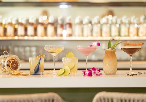 A bar counter with various cocktails and garnishes, including lime slices, orchids, and dried fruits, in front of a selection of bottles.